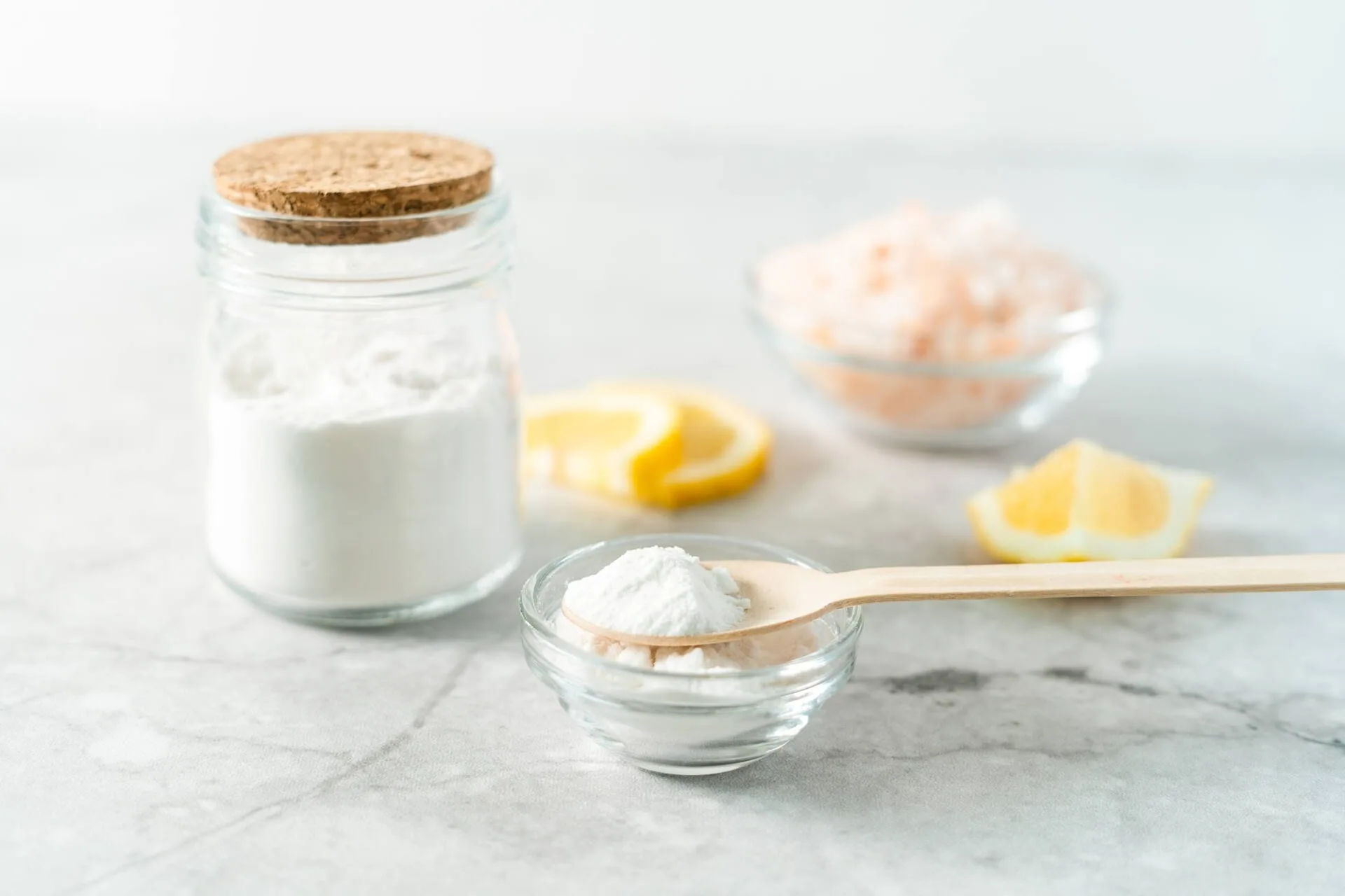A glass jar containing baking soda next to two glass bowls with baking soda, Himalayan Pink Salt and some slices of fresh lemon