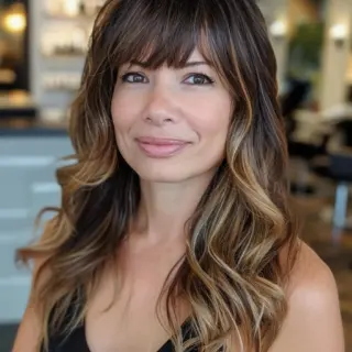 Medium-length waves showcase a striking blend of dark roots melting into golden-brown highlights. Piece-y bangs frame bright eyes while effortlessly connecting to flowing layers. Subtle waves throughout add volume and texture, with longer layers in front creating a beautiful frame that extends past shoulders.