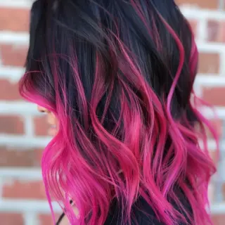 Shoulder-length hair displays an intense black to hot pink ombré effect. Pink sections start subtly at the crown and intensify dramatically through the lengths. Soft waves bring out the multi-dimensional color blend while showing off the sharp contrast between dark and bright shades.